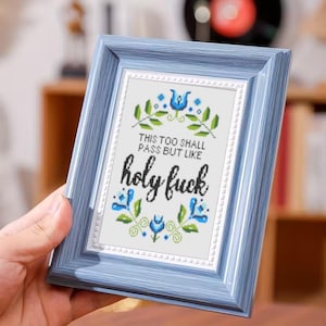 Puede incluir: Un bordado de punto de cruz enmarcado con un marco de madera azul. El bordado presenta el texto "THIS TOO SHALL PASS BUT LIKE holy fuck" rodeado de diseños florales azules y hojas verdes. El marco es de color azul claro.