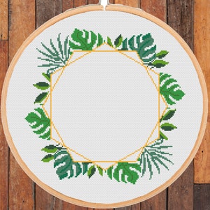 Op de afbeelding: Een ronde borduurring met een witte stoffen achtergrond en een groen bladerig bloemmotief met een gouden geometrisch frame.