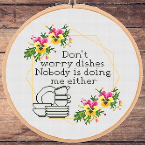 Puede incluir: Un aro de bordado blanco con un diseño de punto de cruz que presenta un borde floral, una pila de platos y el texto "No te preocupes por los platos Nadie los está lavando tampoco".