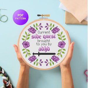 Puede incluir: Un bastidor de bordado de punto de cruz con el texto "Current side quest brought to you by abhd" rodeado por una corona floral. El diseño incluye flores moradas y hojas verdes. Un círculo morado con "PDF PATTERN" es visible.