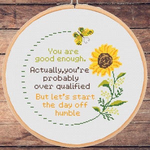 May include: A white embroidery hoop with a cross-stitch design featuring a sunflower, a butterfly, and the text "You are good enough. Actually, you're probably over qualified But let's start the day off humble."