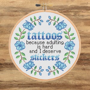 Peut inclure: Un cercle de broderie au point de croix avec le texte "tattoos because adulting is hard and I deserve stickers." Le texte est en bleu, entouré de fleurs bleues et de feuilles vertes. Le cercle est en bois et posé sur une surface en bois.