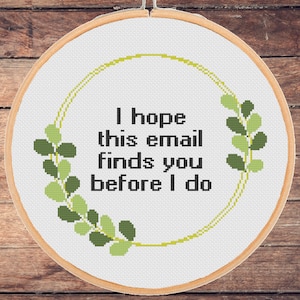 May include: A cross-stitch hoop with the text "I hope this email finds you before I do" in black. The text is surrounded by a green and yellow floral design. The hoop is set against a wooden background.