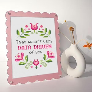 May include: A pink framed cross-stitch with the text "That wasn't very DATA DRIVEN of you" surrounded by floral embroidery. A white vase with dried flowers sits to the right of the frame. The frame has a scalloped edge.