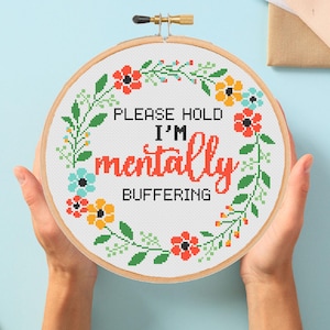 Op de afbeelding: Een borduurring met kruissteek met de tekst "PLEASE HOLD I'M mentally BUFFERING". Het ontwerp toont een bloemenkrans met rode, oranje en blauwe bloemen en groene bladeren. De ring wordt door twee handen vastgehouden.