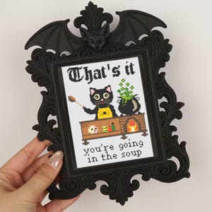Könnte beinhalten: Schwarz gerahmtes Kreuzstich-Kunstwerk mit einer schwarzen Katze in einer gelben Schürze, die einen Kessel umrührt. Der Text lautet: "That's it, you're going in the soup." Der Rahmen hat eine Fledermaus und aufwendige Details, ideal für Halloween.