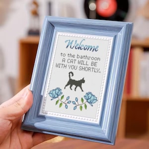 Könnte beinhalten: Ein gerahmtes Kreuzstichbild mit blauem Holzrahmen. Der Text lautet: „Willkommen im Badezimmer, eine Katze wird bald bei Ihnen sein.“ Eine schwarze Katze und blaue Blumenmotive sind ebenfalls abgebildet.