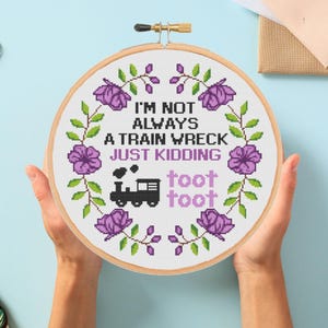 May include: A cross-stitch embroidery hoop with the text "I'M NOT ALWAYS A TRAIN WRECK JUST KIDDING toot toot". The design features a black train, purple flowers, and green leaves, all set against a white background.