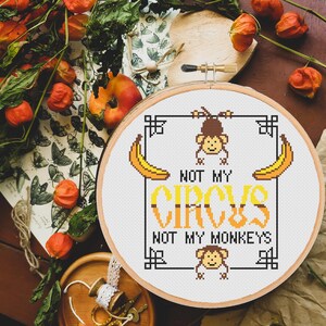May include: A white embroidery hoop with a cross-stitch design that reads "Not my circus, not my monkeys". The design features two monkeys hanging from a bar, two bananas, and a black border.