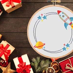 May include: A white embroidery hoop with a blue and white rocket ship, a yellow planet with a red ring, and blue stars on a white background.
