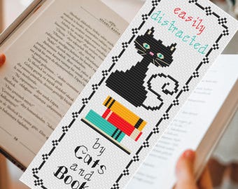 May include: A rectangular cross-stitch bookmark featuring a black cat with green eyes sitting on a stack of red and yellow books. The text reads "easily distracted by Cats and Books" in black and red lettering, all enclosed in a black geometric border.