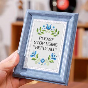 以下が含まれることがあります： 青い木製フレームに入ったクロスステッチ。クロスステッチには、青と緑の花柄に囲まれた黒い文字で「PLEASE STOP USING 'REPLY ALL'」と書かれています。フレームは手で持たれています。