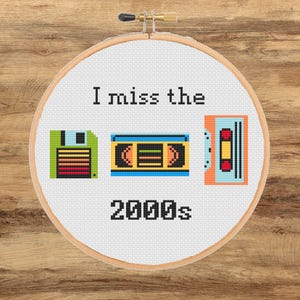 Può includere: Un telaio da ricamo circolare in legno contenente tessuto bianco con un disegno a punto croce. Il testo recita I miss the 2000s, accompagnato da icone pixelate di un floppy disk verde, una videocassetta VHS blu e gialla e una cassetta arancione.