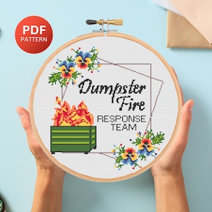May include: A cross-stitch embroidery hoop with the words "Dumpster Fire Response Team" and a graphic of a burning dumpster. The design includes floral accents and is held by two hands. A red circle with "PDF PATTERN" is in the upper left.