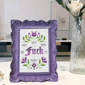 May include: A framed cross-stitch with the text "Off Him Her Fuck It Me That" in a lilac frame. The cross-stitch features floral and leaf designs in green, purple, and white. The frame is a light purple colour with ornate detailing.
