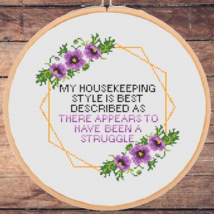May include: A white embroidery hoop with a cross-stitch design featuring purple flowers and a gold geometric frame. The text reads "My housekeeping style is best described as there appears to have been a struggle."