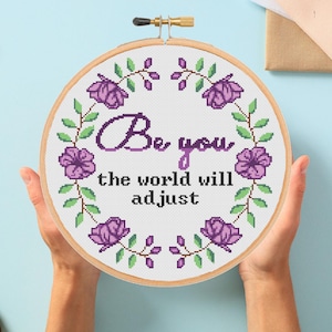 Puede incluir: Un bastidor de bordado de punto de cruz con las palabras "Be you, the world will adjust" en morado. El diseño incluye flores moradas y hojas verdes alrededor del texto. El bastidor es sostenido por manos sobre un fondo azul claro.