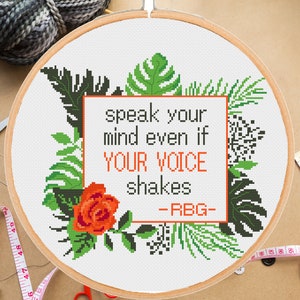以下が含まれることがあります： 白い刺繍枠に、クロスステッチで「speak your mind even if YOUR VOICE shakes -RBG-」という引用が刺繍されています。デザインは緑の葉と赤いバラで囲まれています。