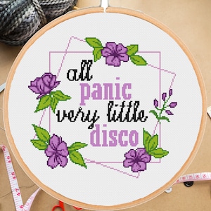 May include: A white embroidery hoop with a cross-stitch design featuring the text "all panic very little disco" surrounded by purple flowers and green leaves.