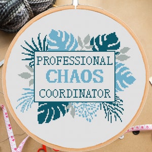 Könnte beinhalten: Stickrahmen mit Kreuzstich-Stickerei mit weißem Hintergrund und blauen und grauen tropischen Blattmotiven. Der Text "Professional Chaos Coordinator" ist in Blau auf einem weißen Rechteck gestickt.