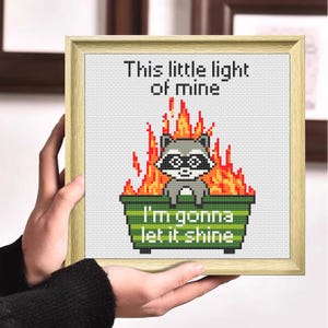 Könnte beinhalten: Kreuzstich-Kunstwerk in einem Holzrahmen. Das Design zeigt einen Waschbären in einem brennenden Müllcontainer mit dem Text "This little light of mine, I'm gonna let it shine."