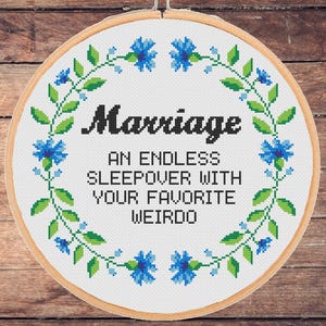 Op de afbeelding: Een witte borduurring met een blauw en groen bloemenontwerp. De tekst "Marriage: An endless sleepover with your favorite weirdo" is geborduurd met zwarte draad.