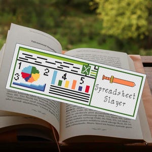May include: A cross-stitched bookmark with a green border, featuring a colorful chart design and the text "Spreadsheet Slayer." The bookmark is placed on an open book with text visible.