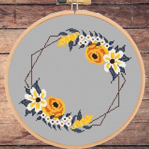 May include: A cross stitch pattern featuring a floral wreath with yellow and white flowers on a gray background. The wreath is framed by a geometric design in brown.