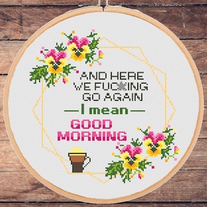 Könnte beinhalten: Ein weißer Stickrahmen mit einem Kreuzstich-Design, das eine florale Umrandung und den Text "And here we f***ing go again - I mean - Good morning" zeigt.
