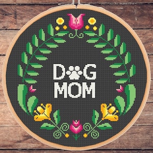 Op de afbeelding: Borduurwerk in een hoepel met een zwarte achtergrond en een groene bloemenkrans. De krans heeft gele bloemen en roze bloemen. De tekst "DOG MOM" staat in wit met een pootprint tussen de woorden.