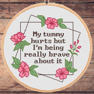 May include: A cross-stitch embroidery hoop with the text "My tummy hurts but I'm being really brave about it." The design includes pink flowers and green leaves. The hoop is set against a wooden background.