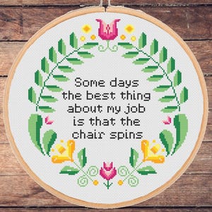 Puede incluir: Un bastidor de punto de cruz con el texto "Some days the best thing about my job is that the chair spins". El diseño incluye una corona de hojas verdes con acentos florales amarillos y rosas. El bastidor está sobre un fondo de madera.