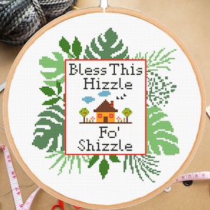May include: A cross-stitch embroidery hoop with a white background and green foliage surrounding a red rectangle with the text "Bless This Hizzle Fo' Shizzle". The rectangle also includes a small yellow house with a brown roof and two green trees.