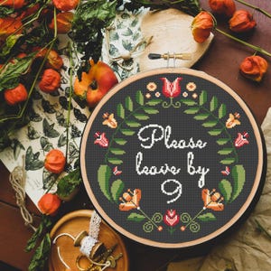 May include: A black embroidery hoop with a floral design and the text "Please leave by 9" in white. The hoop is surrounded by a wooden frame.