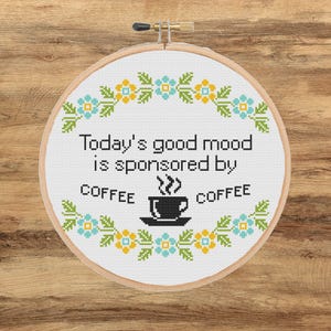 Puede incluir: Un bastidor de bordado circular de madera que contiene tela blanca con un diseño de punto de cruz. El texto dice Today's good mood is sponsored by COFFEE, con un icono de taza de café negra y un borde floral de flores azul claro y amarillas.