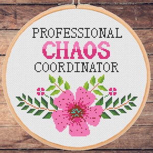 May include: A white embroidery hoop with the text "PROFESSIONAL CHAOS COORDINATOR" in black lettering. A pink flower with green leaves is embroidered below the text.