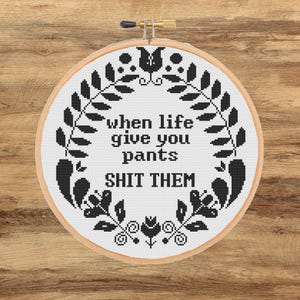 Peut inclure: Un cercle de broderie au point de croix avec l'inscription "when life give you pants SHIT THEM" en fil noir. Le texte est entouré d'une couronne florale noire sur fond blanc. Le cercle est en bois et possède un fermoir doré.