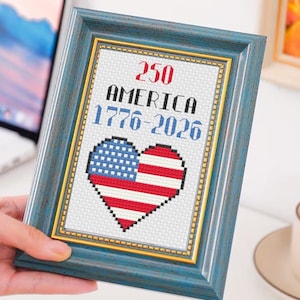 May include: A framed cross-stitch piece with a patriotic theme. The design features a heart-shaped American flag and the text "250 AMERICA 1776-2026" in red and blue. The frame is a distressed blue with a gold inner border.