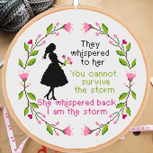 Pu&ograve; includere: Un cerchio da ricamo bianco con un disegno a punto croce che presenta una silhouette di una donna che tiene dei fiori. Il disegno &egrave; circondato da una ghirlanda di foglie verdi e fiori rosa. Il testo dice "They whispered to her You cannot survive the storm She whispered back I am the storm."