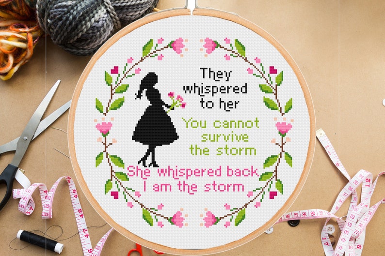 Pu&ograve; includere: Un cerchio da ricamo bianco con un disegno a punto croce che presenta una silhouette di una donna che tiene dei fiori. Il disegno &egrave; circondato da una ghirlanda di foglie verdi e fiori rosa. Il testo dice "They whispered to her You cannot survive the storm She whispered back I am the storm."