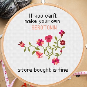 Könnte beinhalten: Ein weißer Stickrahmen mit dem Text "Wenn du dein eigenes SEROTONIN nicht herstellen kannst, ist gekauftes in Ordnung" und einem rosa-grünen Blumendesign, das die chemische Struktur von Serotonin darstellt.