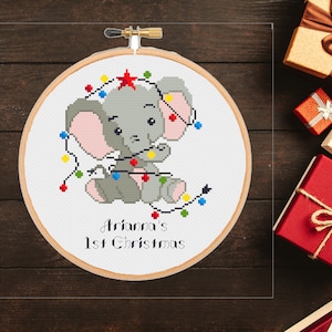May include: A white embroidery hoop with a cross-stitch design of a gray elephant with pink ears holding colorful Christmas lights. The text "Ariana's 1st Christmas" is written below the elephant.