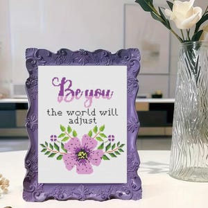 May include: A framed cross-stitch artwork with the words "Be you, the world will adjust" above a floral design. The frame is a decorative purple color.