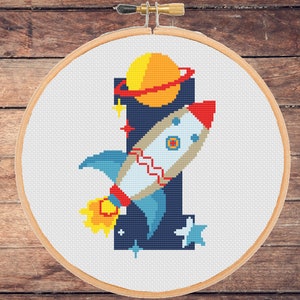 Op de afbeelding: Een kruissteekpatroon met een raket die door de ruimte vliegt met een planeet en sterren. De raket is blauw, wit en rood. De planeet is oranje en geel. De sterren zijn wit. De achtergrond is donkerblauw.