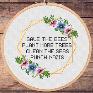 May include: A cross-stitch embroidery in a wooden hoop. The design features a geometric gold frame with the text "SAVE THE BEES PLANT MORE TREES CLEAN THE SEAS PUNCH NAZIS". Floral accents in pink, blue, and green surround the text.