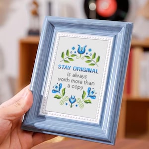 May include: A blue framed picture with the text "STAY ORIGINAL is always worth more than a copy". The picture has a white background with blue and green floral embroidery.