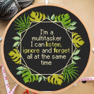 Puede incluir: Bordado de punto de cruz en un aro de madera con el texto "I'm a multitasker I can listen, ignore and forget all at the same time." El diseño presenta un fondo negro con hojas tropicales verdes y un marco geométrico.
