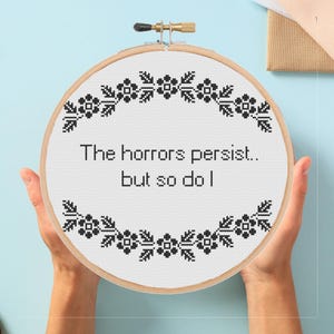 May include: A cross-stitch embroidery hoop with the text "The horrors persist.. but so do I." The design features a floral border in black thread. The hoop is made of wood and is held by two hands.