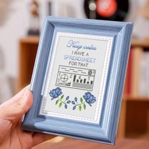 May include: A framed cross-stitch with a pale blue frame. The cross-stitch reads "Keep calm I have a spreadsheet for that" in blue script. Below the text is a graphic of a spreadsheet and blue floral accents.