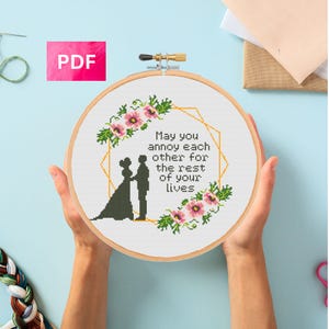 Puede incluir: Un bastidor de bordado de punto de cruz con el texto "May you annoy each other for the rest of your lives". El diseño presenta una silueta de novios, flores rosas y hojas verdes. Un icono PDF está en la parte superior izquierda.